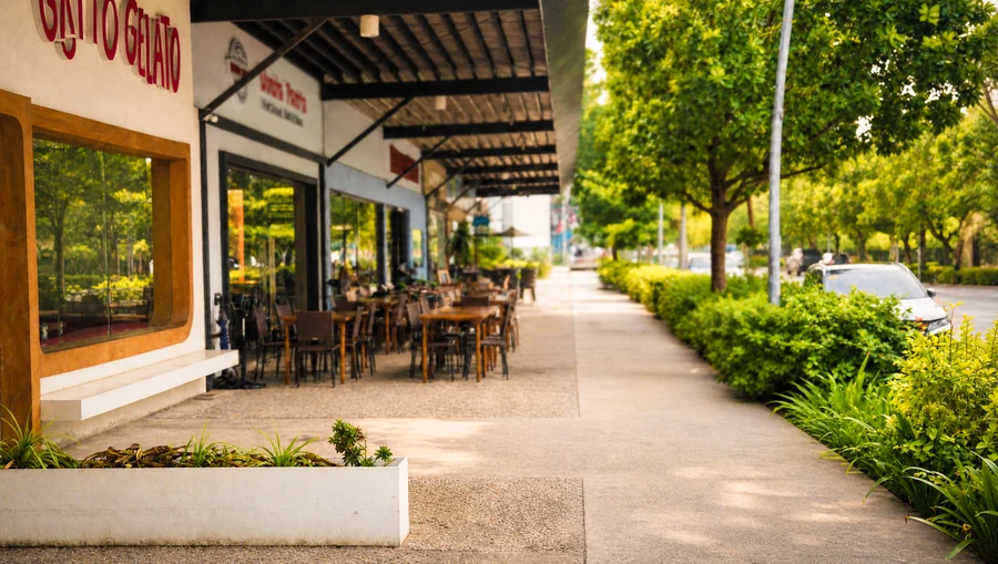 The sunny covered walkway outside Gatto Gelato on The Riverside Strip, outdoor tables under the awning and a tree-lined sidewalk